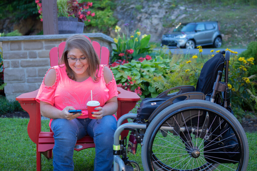 Dans un parc, à la tombé du jour, une jeune femme est assise dans une chaise de jardin rouge, elle tient un café et elle joue avec son téléphone. Elle a une tenue d'été, un haut rose et un jeans, à coté d'elle se tient son fauteuil roulant manuel.