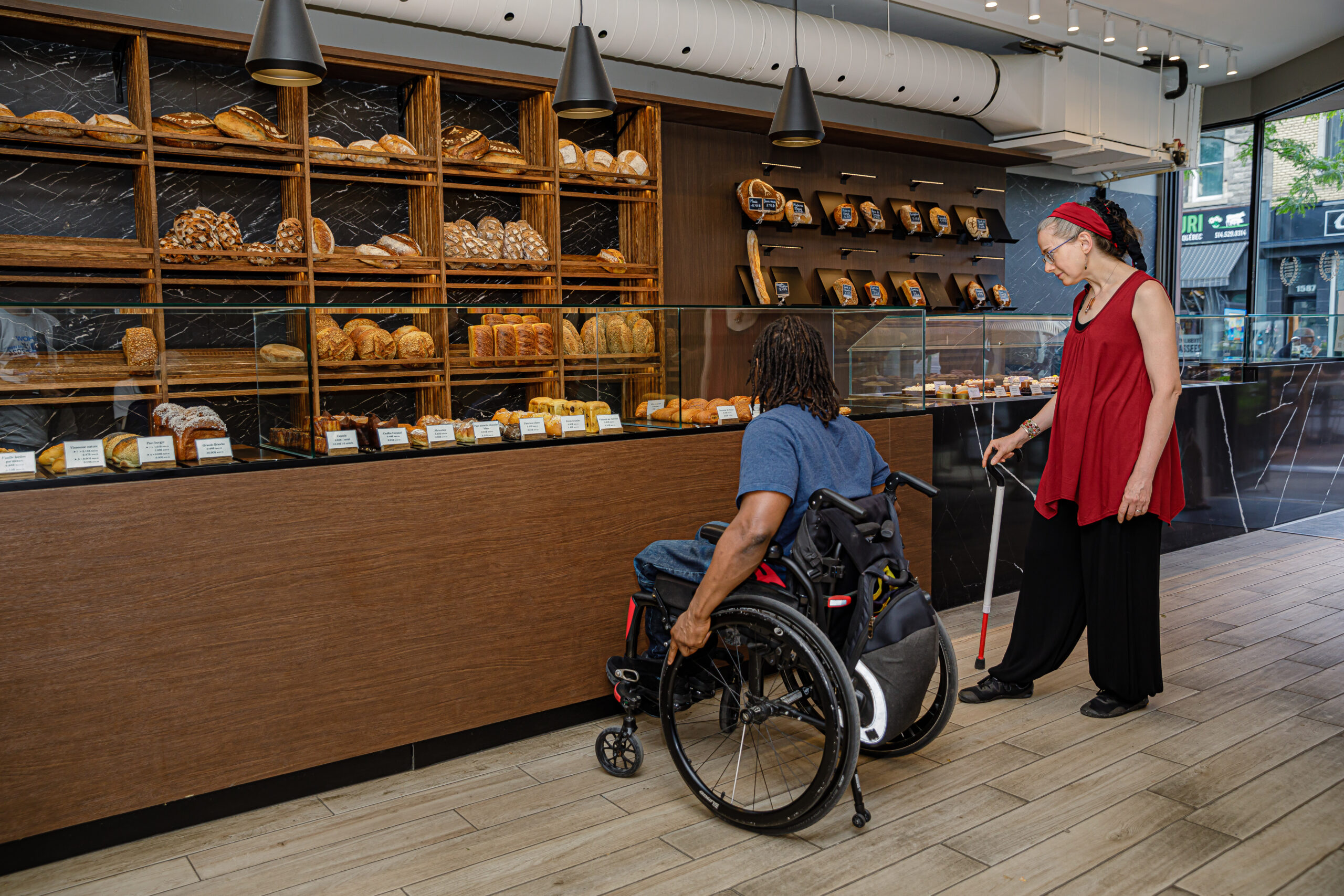 une femme avec une canne blanche et un homme en fauteuil manuel regarde les produits dans la vitirine d'une boulangerie